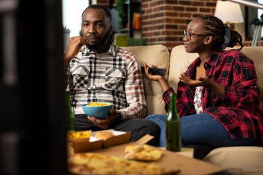 Genç çift rahat oturma odasındaki kanepede dinleniyor. Siyahi kadın uzaktan kumandayı tutuyor ve erkek partneriyle konuşuyor. O da televizyon seyredip abur cubur yiyor. Akşamları evde takılıyorum..