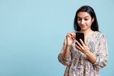 Stüdyoda cep telefonu ekranında gezinen neşeli, şaşkın kız mavi arka planda online alışveriş yapıyor. İnternet sitelerine girip hayret içinde haykıran sıradan bir kadının portresi.