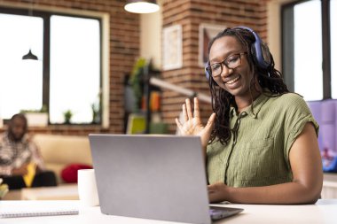 Neşeli Afro-Amerikalı kadın kulaklık takıyor, sanal toplantı sırasında dizüstü bilgisayarında el sallıyor. İçerde oturan siyahi kadın girişimci, uzaktan video konferansında müşterileri selamlıyor.