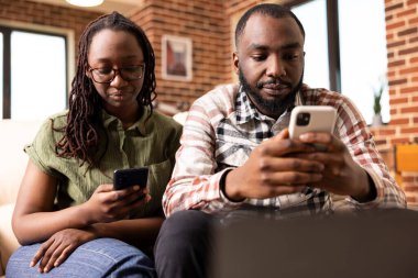 African american husband and wife sitting on couch, each focused on their mobile phones. Young black couple browsing social media and texting, enjoying digital lifestyle in brick wall apartment.
