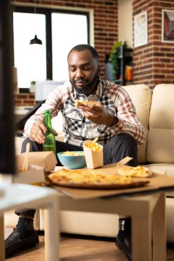 Portrait of bearded guy seated on couch, having pizza slice and cold drink during casual weekend. Black man watching television and enjoying quiet afternoon alone in brick wall apartment.