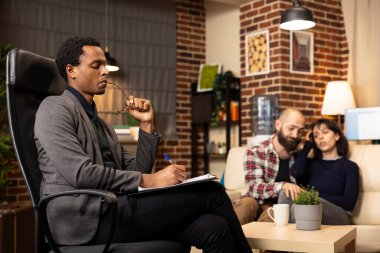 Odaklanmış erkek psikiyatrist, danışmanlık sırasında beyaz bir çifti dikkatle dinlerken gözlük ısırıyor ve panoya yazıyor. Profesyonel terapist devam eden terapi seansında..