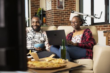 Kadın dizüstü bilgisayar kullanırken, bir kase cipsi olan Afro-Amerikalı bir adam televizyonda dizi izliyor. Siyahi erkek arkadaş ve kız arkadaş yemek ve dijital eğlenceyle evde dinleniyor..