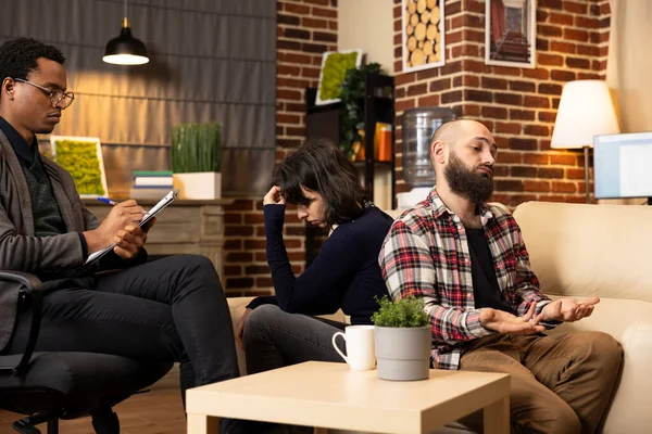 Professional family psychologist sits in chair, listening to clients seated back to back during marriage counseling. African American therapist analyzing relationship issues during couple therapy.