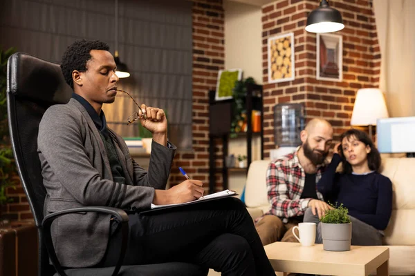 Odaklanmış erkek psikiyatrist, danışmanlık sırasında beyaz bir çifti dikkatle dinlerken gözlük ısırıyor ve panoya yazıyor. Profesyonel terapist devam eden terapi seansında..