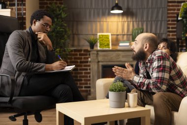 Diverse individuals participating in ongoing therapy session. Black counselor listening attentively and taking notes on clipboard as white male patient discusses relationship issues.