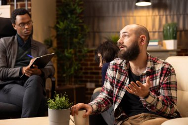 Professional counselor listening to white couple seated back to back during marriage counseling. Therapist takes notes as man shares relationship challenges, analyzing issues to guide couple therapy.
