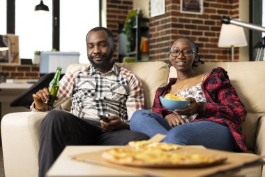 Genç çift evde dinleniyor, hafta sonları pizza, bira ve patates kızartmasıyla eğleniyor. Siyah kadın ve erkek rahat bir koltukta oturmuş, oturma odasında film ya da spor maçı izliyorlar..