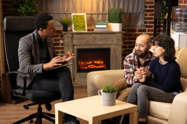 Young couple sits closely on couch holding hands during marriage counseling session. Professional psychologist holds glasses while guiding open conversation, offering support and relationship advice.