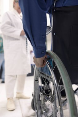 Engelli beyaz erkek hasta tıbbi randevu için kliniğe geliyor. Yakın plan, iyileşme ve sağlık planlaması sırasında gücü ve dayanıklılığı sembolize eden tekerlekli sandalyeli bir el olduğunu gösteriyor..