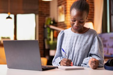 Kapalı alanda çalışan kadın serbest çalışan, dizüstü bilgisayardaki satış performansını inceleyen ve not alan biri. Rapor ve organizasyonel görevleri yönetirken not defterine fikirler yazan uzaktan çalışan.