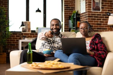Black boyfriend and girlfriend enjoying quiet time in cozy living room. Bearded man enjoying snacks and TV while woman stays focused on laptop, balancing work or study during relaxed weekend downtime.