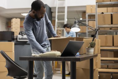 Black male staff verifying custom orders logistics and product handling, working at a fulfillment center workstation. Packing goods in the storage room of growing local shipping startup.
