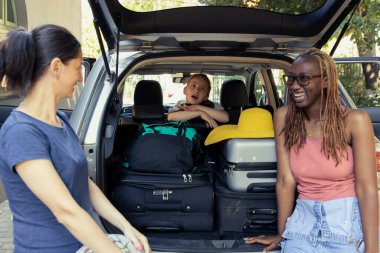 On a bright summer day, young family and close friends load their car trunk with luggage. Their road trip begins with laughter, teamwork and excitement for a seaside escape full of leisure.