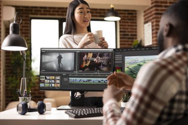 African american videographer pausing remote work to listen to asian girlfriend holding coffee cup in cozy brick wall living room. Supportive wife speaking with husband during film post production.