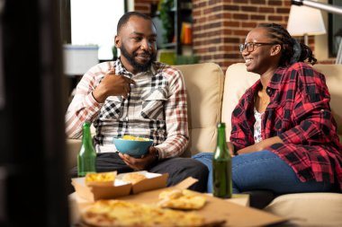Mutlu siyah çift kanepede kaliteli zaman geçiriyor, yemek yiyor ve rahat bir dairede sohbet ediyor. Modern tuğla duvarlı oturma odasında yemek ve içeceklerle rahatça takılabiliriz..