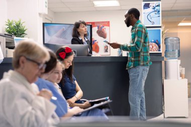 Siyahi adam tıbbi atamadan önce hastane resepsiyonistinden tavsiye istiyor. Resepsiyondaki beyaz kadın klinikte bekleyen erkeğe sağlık sigortası bilgisi ve el ilanı veriyor.