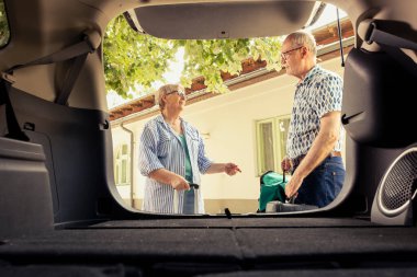 Smiling senior couple packing a suitcase into their automobile trunk, preparing for enjoyable road trip through mountain or countryside. Embracing the fun of retired life and freedom.