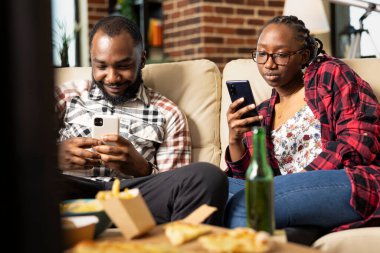 Afro-Amerikalı kadın ve erkek koltukta rahat bir şekilde otururlar, her biri akıllı telefonlarına odaklanırlar. Haftasonu atıştırmalıklarla takılmak, teknolojiyle kaynaşmak, modern bir daire..