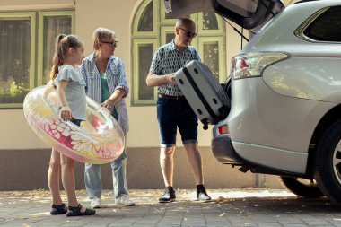 Retired couple and their niece smiles as they load travel bags into the vehicle. Seaside road trip getaway represents relaxed holiday and happy summertime leisure activity, beach fun.