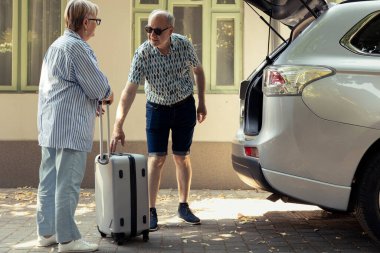 Retired people loading the car with baggage and enjoying free time, feeling excited as they prepare for relaxing vacation and road trip. Elderly man and woman enjoys travelling together.