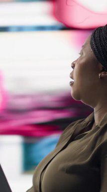 Vertical video Woman uses laptop, enjoying leisure time with content on video streaming services. African american person watches media VOD broadcasts on notebook in front of digital backdrop with