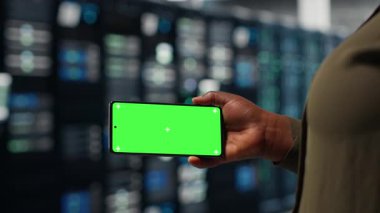 Software developer using app on mockup phone to check server hub energy consumption. African american woman using green screen smartphone in data center to do gear checkup, camera B