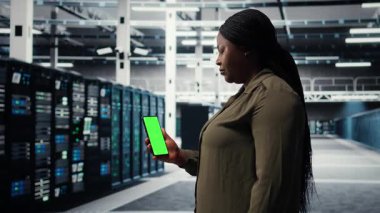 Software developer using app on mockup phone to check server hub energy consumption. African american woman using green screen smartphone in data center to do gear checkup, camera A