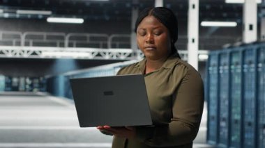 Technician in server room using laptop to upgrade rigs for increase in clients workload capacity. African american woman in data center configures equipment, improving processing speed, camera B