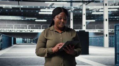 Data center programmer monitors data center metrics on tablet to enhance gear operational effectiveness. African american employee in server hub using analytics to predict performance, camera A