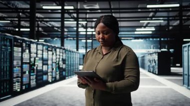 Data center software developer monitors data center metrics on tablet to enhance operational effectiveness. Woman in server room using app device analytics to predict performance, camera A