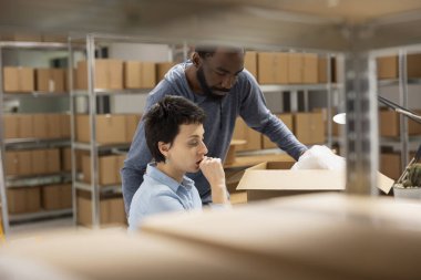 İki farklı işçi, istiflenmiş kutular ve organize ürün envanterleriyle çevrili bir depoda, verimli küçük işletme operasyonları içeren sevkiyat lojistiğini tartışıyor..