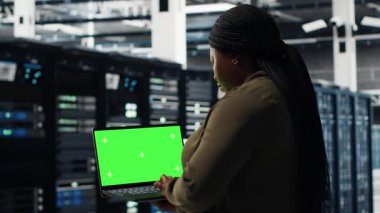 Server room admin uses green screen laptop to upgrade mainframes for increase in workload capacity. Data center worker configures gear with mockup notebook, improving processing speed, camera B