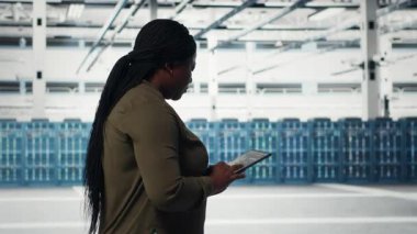 Technician walking in data center with tablet, checking hardware for system stability. Worker overseeing server farm with device, configuring infrastructure rigs, camera B