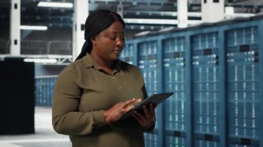 Data center programmer monitors data center metrics on tablet to enhance gear operational effectiveness. African american employee in server hub using analytics to predict performance, camera B