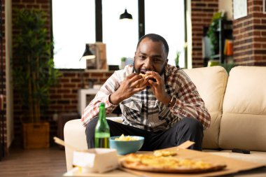 Black male freelancer taking break from work, sitting on couch and enjoying slice of pizza. Relaxed african american man watching movie or show at home with takeaway meal in hand.