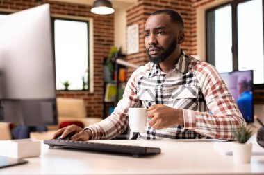 Ofiste serbest çalışan siyahi bir adam endişeli bir ifadeyle masaüstü monitörüne bakıyor, şirket belgelerini inceliyor. Elinde kahve fincanı tutan ve mali verileri kontrol eden genç bir girişimci.