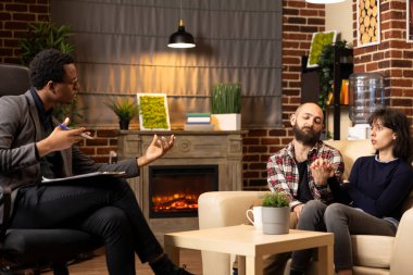 Terapist çiftin duygusal refahını tartışırken kocanın yanında oturan kadın müşteri, terapi seansı sırasında endişeli görünüyor. Siyahi erkek danışman terapide eşlere danışmanlık yapıyor.