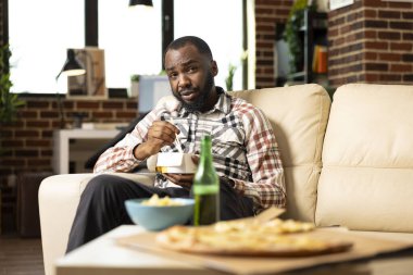 Young bearded man enjoys relaxed dinner at home, eating takeaway pasta while watching movie. Bearded guy seated on sofa in cozy brick wall apartment, embracing solo comfort and leisure time.