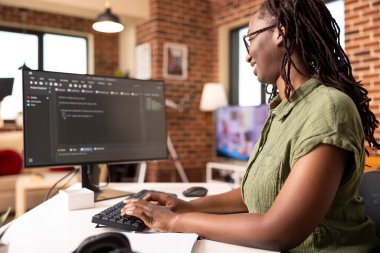 Black female software developer codes securely from home office, using computer to review programming scripts. African american IT freelancer working on tech solutions in modern apartment.