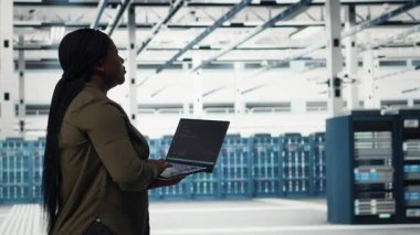 Sistem istikrarı için yazılım güncellemelerini yönetmek için dizüstü bilgisayar kullanan bir veri merkezi çalışanı. Sunucu odasında Afro-Amerikalı bir kadın var. Optimum performans için altyapıyı ayarlıyor. Kamera B.