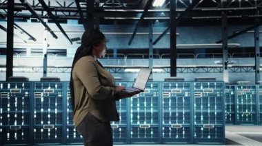 Engineer walks in server hub, using laptop to upgrade rigs for increase in clients workload capacity. Data center employee configures equipment with notebook, improving processing speed, camera A