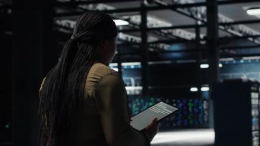 Engineer in data center using tablet to deploy secure server architecture. African american woman in server hub using device to do rollout of cybersecurity framework, camera B