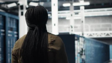 Woman walking through data center server rows, checking equipment for system stability. IT professional overseeing server farm, monitoring infrastructure mainframes, camera B