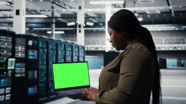 Server room admin uses green screen laptop to upgrade mainframes for increase in workload capacity. Data center worker configures gear with mockup notebook, improving processing speed, camera A