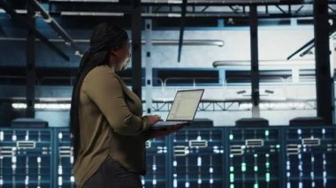 Software developer in data center using laptop to coordinate deployment of new server architecture. Server hub IT professional using notebook to do rollout of cybersecurity framework, camera B