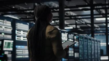 Data center system administrator monitors data center metrics on tablet to enhance operational effectiveness. IT expert in server room using analytics program to predict performance, camera B