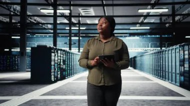 Data center engineer monitors data center metrics on tablet to enhance rigs operational effectiveness. Worker walks through server farm rows, using analytics on device to predict performance, camera A