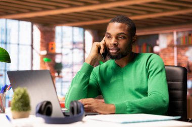 Freelancer in home office having important phone call with business partner using smartphone. African american person sitting at desk discussing company matters during telephone call