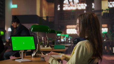 İlkokul öğrencisi kütüphanede masada oturuyor ve tabletin yanında kitap okuyor, gelecek dersler için notlar ve fikirler hazırlıyor. Alim kız ödev olarak dijital cihaz kullanıyor. Kamera A.